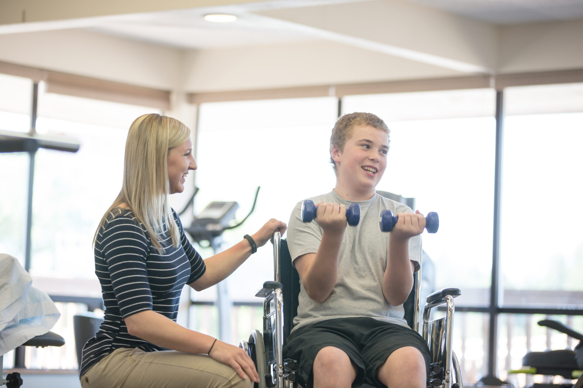 Physical therapist works with young adolescent patient in wheelchair to help with recovery. He is lifting small weights.